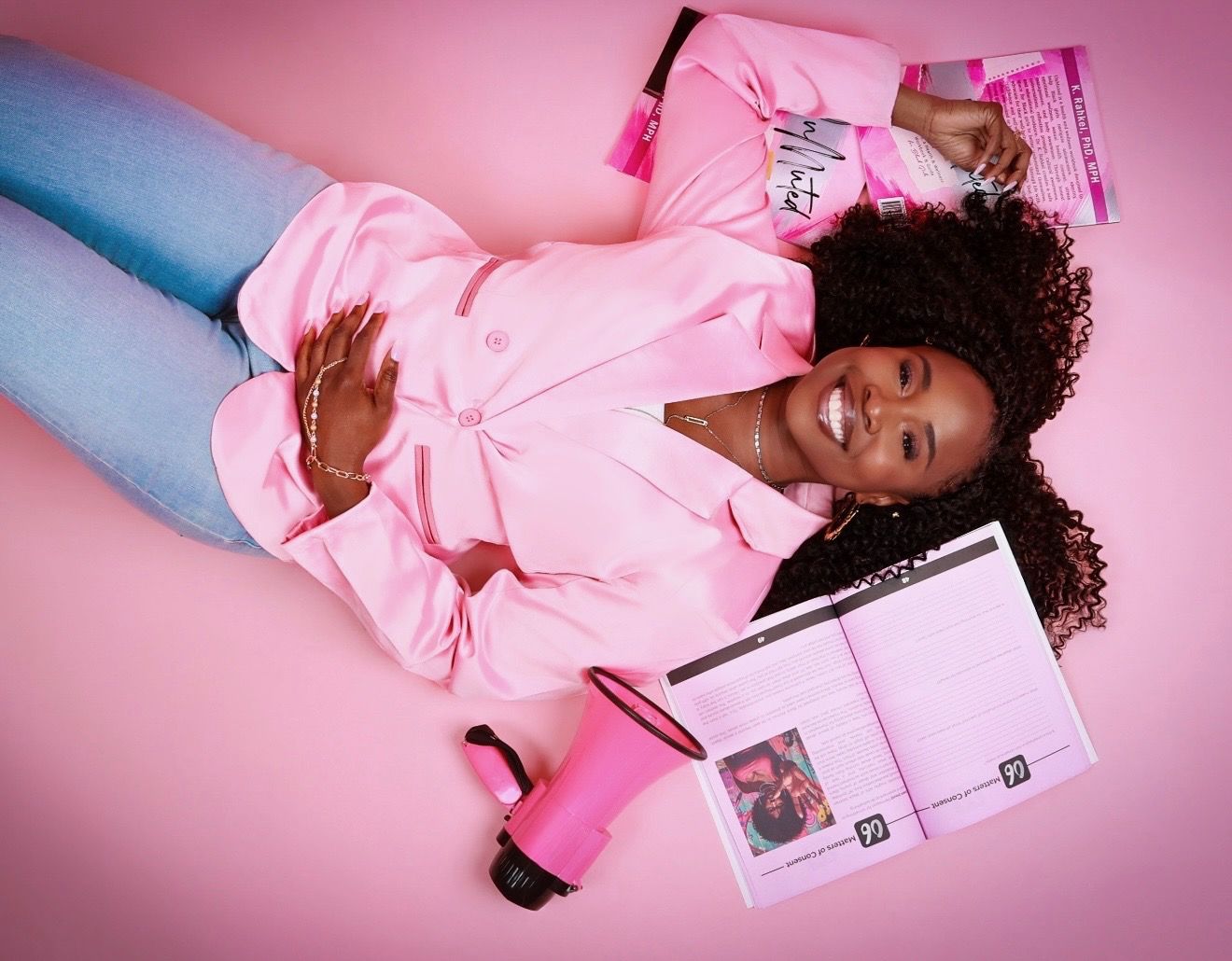 Dr. K. Rahkel lying on a pink floor surrounded by UnMuted books open to Matters of Consent, with a pink megaphone